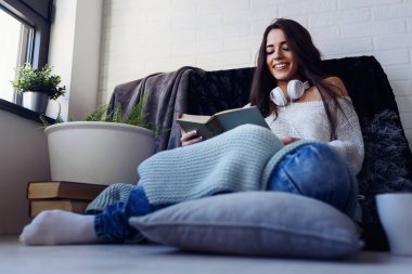 Güzel genç bir kadın kitap okuyor, modern apartmanın penceresinin önünde eğleniyor..