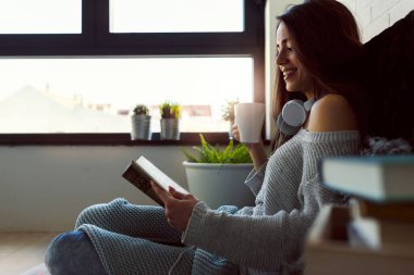 Güzel genç bir kadın kitap okuyor, modern apartmanın penceresinin önünde eğleniyor..