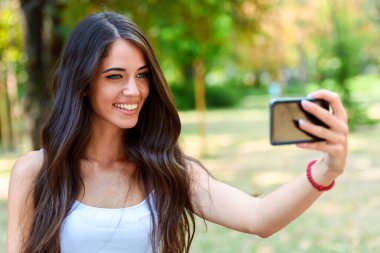 Genç ve güzel bir kadın parkta cep telefonuyla selfie çekiyor. 
