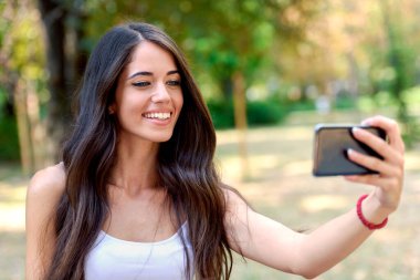 Genç ve güzel bir kadın parkta cep telefonuyla selfie çekiyor. 