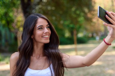 Genç ve güzel bir kadın parkta cep telefonuyla selfie çekiyor. 