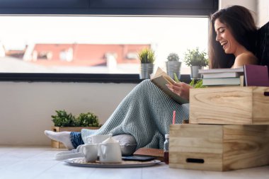 Güzel genç bir kadın kitap okuyor, modern apartmanın penceresinin önünde eğleniyor..