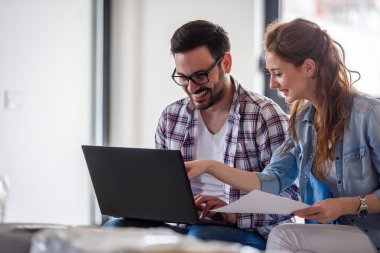 Mutlu genç çift dizüstü bilgisayar kullanıyor, yeni evine taşınıyor.