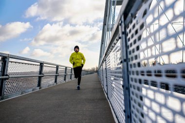 Çekici formda bir adam büyük ve modern bir köprü boyunca hızla koşuyor. Egzersiz, koşu, spor, kış. Erkek sporcu.