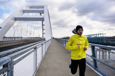 Çekici formda bir adam büyük ve modern bir köprü boyunca hızla koşuyor. Egzersiz, koşu, spor, kış. Erkek sporcu.