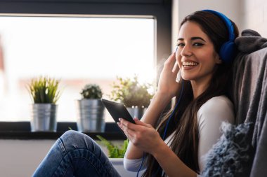 Kulaklıklı genç bir kadın kanepede oturuyor ve tablette müzik dinliyor.