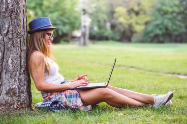 Güzel, genç bir kız öğrenci, dizüstü bilgisayarıyla parktaki yeşil çimlerin üzerinde oturuyor. Uzaklık öğrenimi