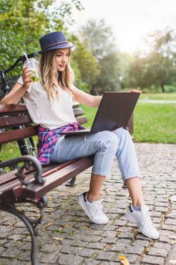 Dizüstü bilgisayarı olan genç bir kadın bankta oturuyor ve bilgisayar kullanıyor.