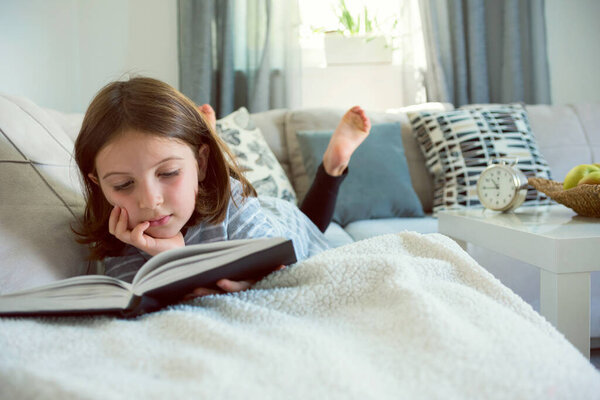 красивая маленькая девочка читает книгу на диване 