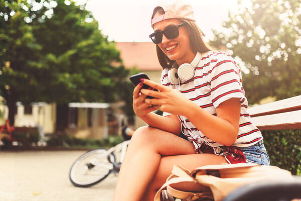 Улыбающаяся девушка в солнечных очках, сидящая летом на скамейке запасных, используя смартфон