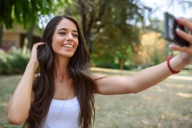 Uzun kahverengi saçlı genç güzel parkta akıllı telefondan selfie çekiyor.