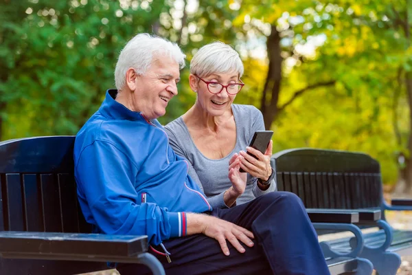 Smiling Senior Active Couple Sitting Bench Looking Smartphone — Stock Photo, Image