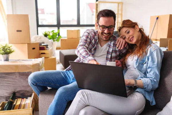 stock image Beautiful young couple sitting among cardboard boxes in their new apartment making plans with laptop