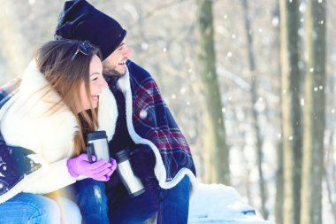 Karla kaplı dağ ormanlarında oturan gülümseyen genç bir çift. Sıcak içecekler içmek, eğlenmek..