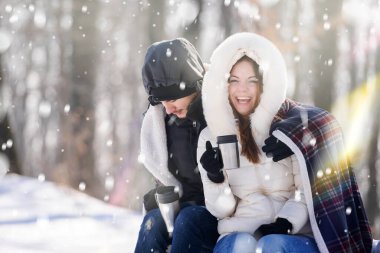 Karla kaplı dağ ormanlarında oturan gülümseyen genç bir çift. Sıcak içecekler içmek, eğlenmek..