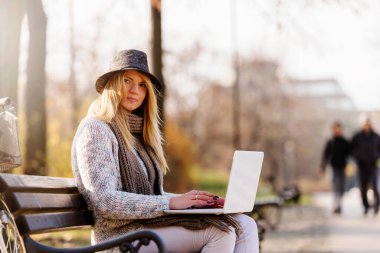 Dizüstü bilgisayarı olan genç bir kadın parkta bankta oturuyor.