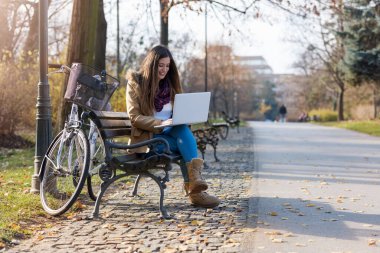 Bisikletli gülümseyen öğrenci kız bankta dizüstü bilgisayarla çalışıyor.