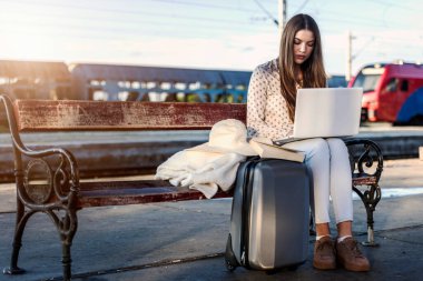 Tren istasyonunda bavullu bir kız dizüstü bilgisayarla çalışıyor.