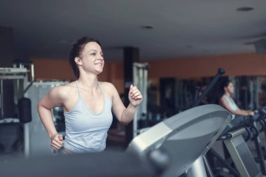 Beautiful cheerful fitness young woman in sportswear running on a treadmill in a fitness center. Sporty beauty at the gym. clipart