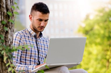 Genç adam parkta dizüstü bilgisayarla çalışıyor. 