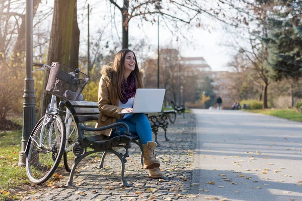 Stock image Smiling student girl with bicycle on park bench, working on laptop