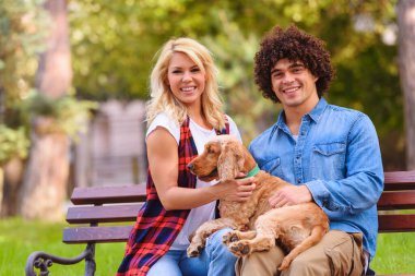 Köpeğiyle parktaki bankta oturan gülümseyen genç çift.