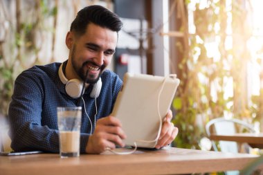 Kulaklıklı genç adam kafede tablet kullanarak oturuyor.