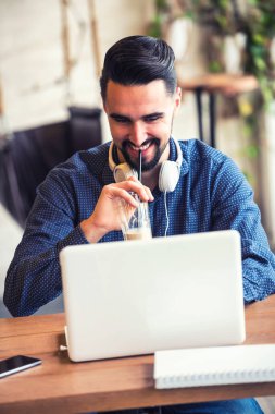 Sakallı neşeli genç adam kahve dükkanında dizüstü bilgisayarla çalışıyor.