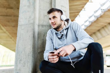 Kulaklıklı yakışıklı bir koşucu köprü altında mola verip müzik dinliyor.