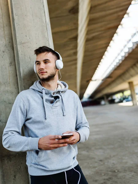 stock image Urban handsome man runner with earphones taking break under bridge listening music