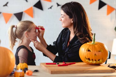 Mutlu anne ve kız Cadılar Bayramı 'na hazırlanıyor, anne ve kız çocukları yüzlerini boyuyor.