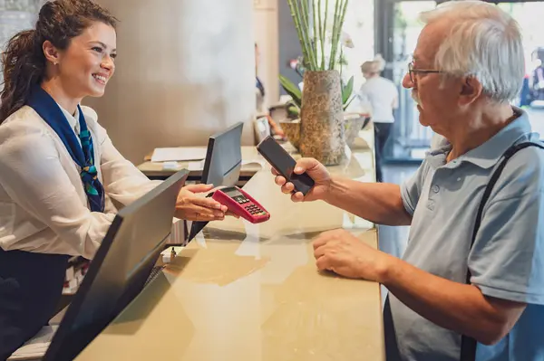 A senior couple at the hotel front desk makes contactless payments with a smartphone. Seniors traveling and using new technologies