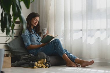 Güzel genç bir kadın kitap okuyor, pencerenin kenarında rahat bir şekilde oturuyor.