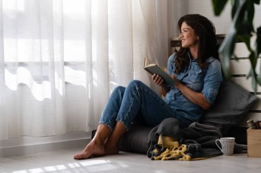 Güzel genç bir kadın kitap okuyor, pencerenin kenarında rahat bir şekilde oturuyor.