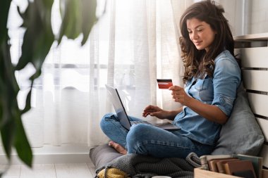 Güzel gülümseyen genç bir kadın evdeki pencerenin kenarında rahat bir şekilde oturuyor, bilgisayarını kullanarak online tatil hediyelerini kredi kartıyla alıyor.