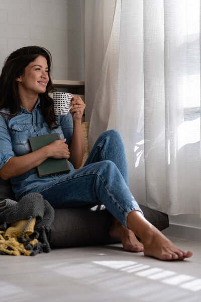 Beautiful smiling young woman sitting at home in cozy window corner, drinking coffee, relaxing, enjoying a morning moment of peace