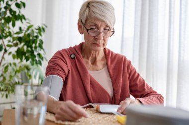 Yaşlı kadın evde tek başına oturmuş, kan basıncını ölçüyor.