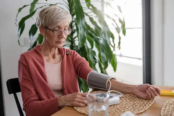 自宅で一人で座り 家庭用デバイスで血圧を測定するシニア女性 — ストック写真
