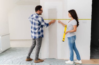 Bir adam ve hamile karısından oluşan genç, güzel ve neşeli bir çift, yeni ev yenileme projesinin bir parçası olarak duvarları ölçüyorlar.