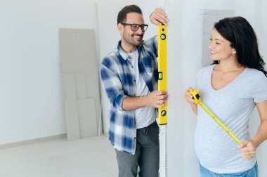 Bir adam ve hamile karısından oluşan genç, güzel ve neşeli bir çift, yeni ev yenileme projesinin bir parçası olarak duvarları ölçüyorlar.