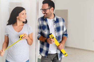 Bir adam ve hamile karısından oluşan genç, güzel ve neşeli bir çift, yeni ev yenileme projesinin bir parçası olarak duvarları ölçüyorlar.