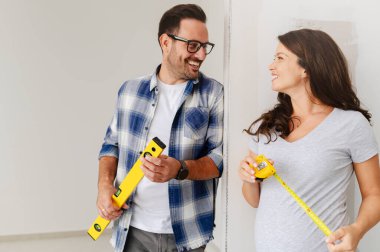 Bir adam ve hamile karısından oluşan genç, güzel ve neşeli bir çift, yeni ev yenileme projesinin bir parçası olarak duvarları ölçüyorlar.