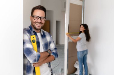 Bir adam ve hamile karısından oluşan genç, güzel ve neşeli bir çift, yeni ev yenileme projesinin bir parçası olarak duvarları ölçüyorlar.