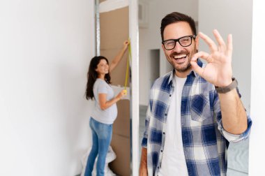 Bir adam ve hamile karısından oluşan genç, güzel ve neşeli bir çift, yeni ev yenileme projesinin bir parçası olarak duvarları ölçüyorlar.