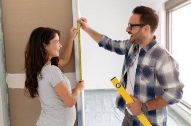 Bir adam ve hamile karısından oluşan genç, güzel ve neşeli bir çift, yeni ev yenileme projesinin bir parçası olarak duvarları ölçüyorlar.