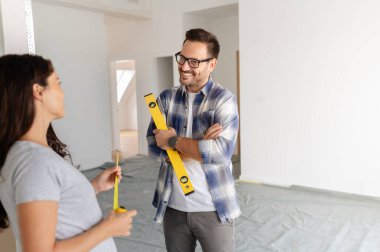 Bir adam ve hamile karısından oluşan genç, güzel ve neşeli bir çift, yeni ev yenileme projesinin bir parçası olarak duvarları ölçüyorlar.