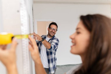 Bir adam ve hamile karısından oluşan genç, güzel ve neşeli bir çift, yeni ev yenileme projesinin bir parçası olarak duvarları ölçüyorlar.