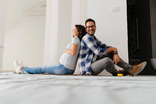 man and his pregnant wife are seating on the floor of an empty house.