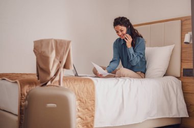 Oteldeki yatağında dizüstü bilgisayarı ve cep telefonu olan genç bir kadın.