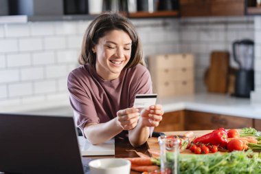 Genç ve çekici bir kadın kredi kartı tutuyor ve online alışverişi için online alışveriş yapıyor. 